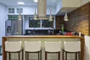 Cozinha americana com bancada, pendentes prateados e cadeiras brancas com altura ideal pendente.