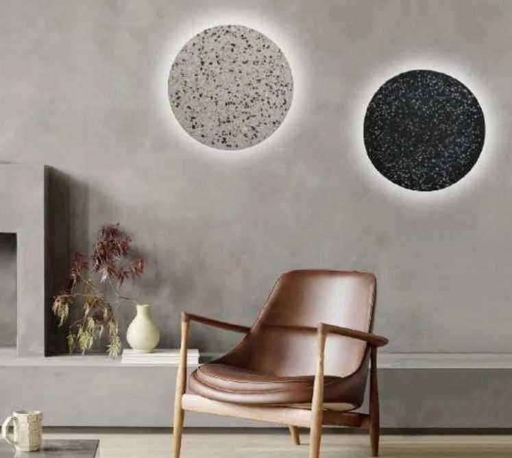 Arandelas circulares com acabamento texturizado, uma em tom claro e outra em tom escuro, montadas em uma parede de cimento queimado. As peças possuem iluminação suave ao redor, criando um efeito de luz difusa que realça o design minimalista.