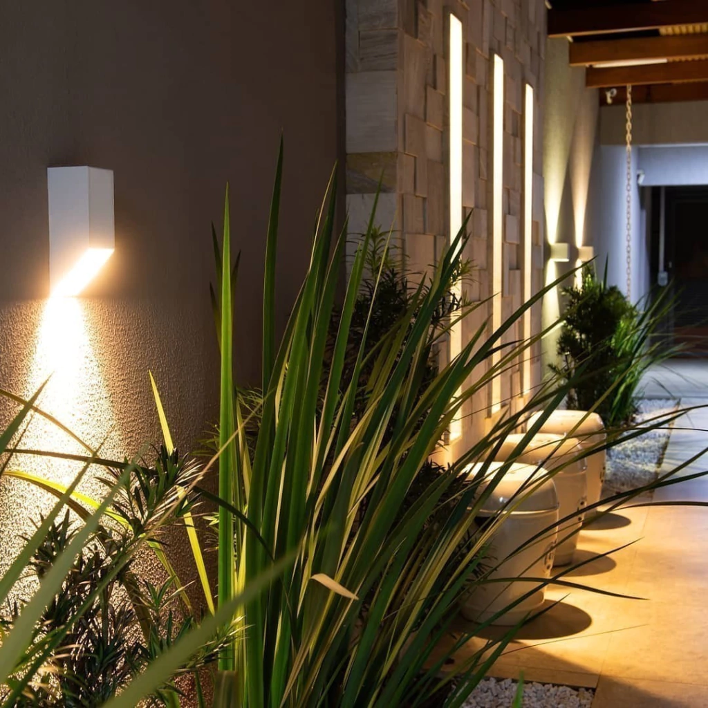 Área externa com arandelas iluminando plantas e paredes texturizadas.