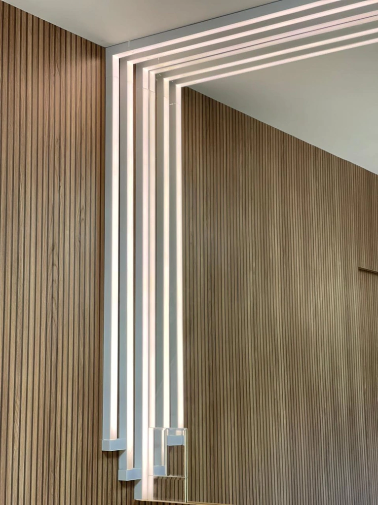 Perfil de LED Nazca instalado em uma parede moderna com acabamento em madeira e sofás amarelos, refletido em espelho. Lustres Gênesis loja online de Lustres e Iluminação Decorativa.