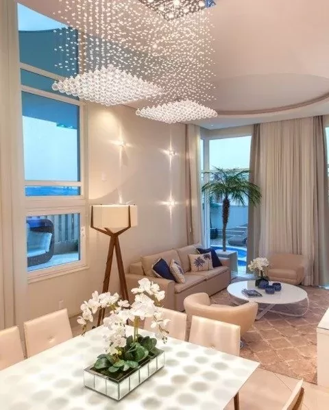 Interior de uma sala de estar moderna com decoração elegante, incluindo um lustre de cristal, sofá bege com almofadas azuis, mesa de centro branca e plantas decorativas. Lustres Gênesis loja online de Lustres e Iluminação Decorativa.