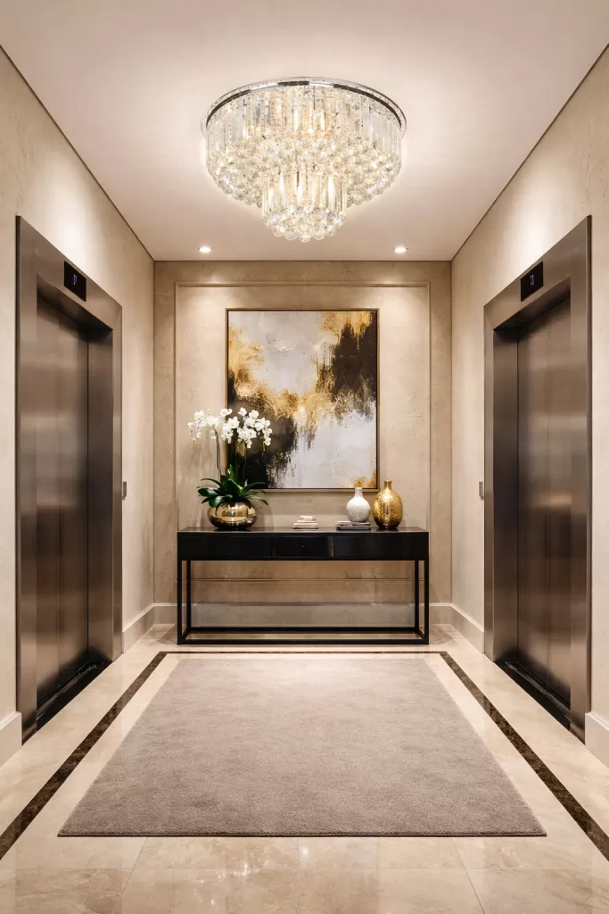 Lustre de cristal elegante instalado no hall de elevador, com design em camadas e cristais lapidados que refletem a luz quente de forma sofisticada. A peça centraliza o ambiente e valoriza o espaço com brilho intenso, criando uma atmosfera luxuosa e acolhedora no lobby.