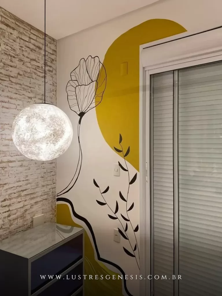 Lustre pendente esférico com efeito lunar e acabamento texturizado branco, suspenso por cabo preto, iluminando o ambiente com luz suave e difusa. A peça se destaca ao lado de uma parede com pintura artística em tons de amarelo, branco e preto, criando um contraste moderno com o revestimento de tijolo aparente e trazendo personalidade ao espaço.