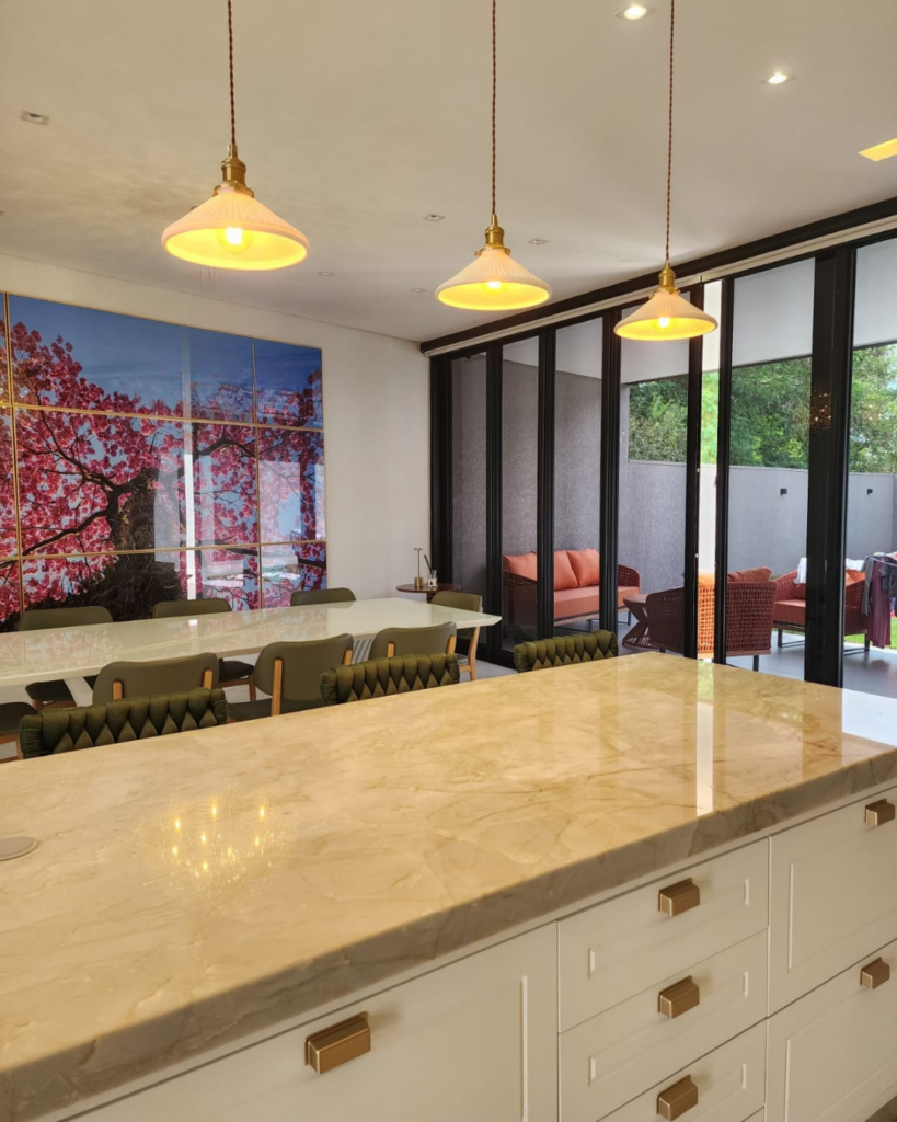 Cozinha gourmet integrada com sala de jantar, iluminada por três pendentes com cúpula branca e detalhe dourado sobre ilha de mármore bege. A iluminação quente valoriza o ambiente moderno, com portas de vidro, varanda externa e mesa de jantar elegante ao fundo.