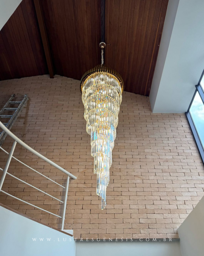 Lustre de cristal para pé direito duplo com design em cascata, composto por múltiplos níveis de tubos transparentes suspensos por base dourada. Instalado sobre escada com parede de tijolos aparentes e teto em madeira, a peça cria um efeito luminoso sofisticado e imponente, ideal para halls residenciais de alto padrão.