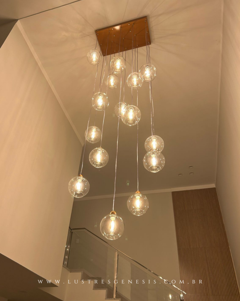 Lustre pendente para pé direito duplo com base retangular amadeirada e múltiplos fios suspensos em diferentes alturas, formando uma composição assimétrica de globos de vidro transparente. As lâmpadas de filamento aparente criam iluminação quente e aconchegante, refletida no painel de vidro ao fundo, ideal para halls e salas com escada e design contemporâneo.