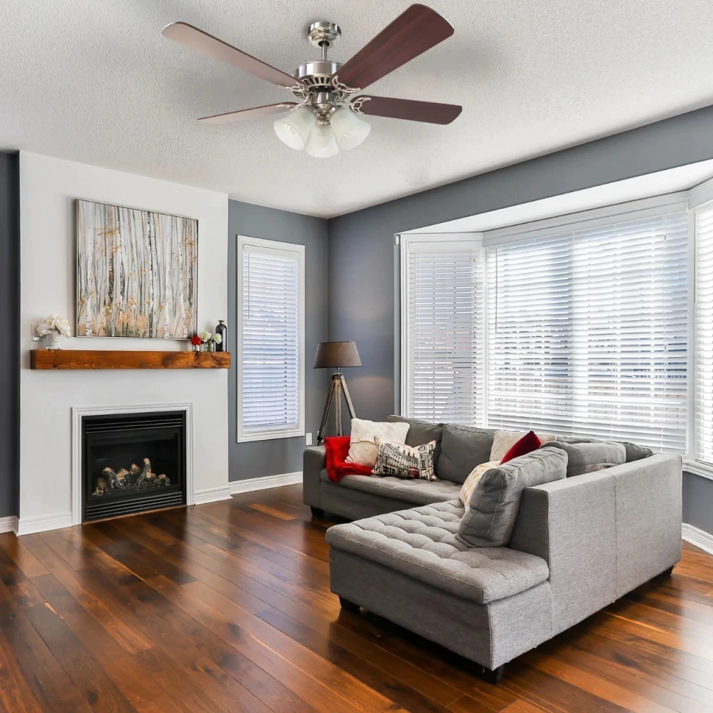 Sala aconchegante com sofá cinza, ventilador de teto de 4 pás e lareira.