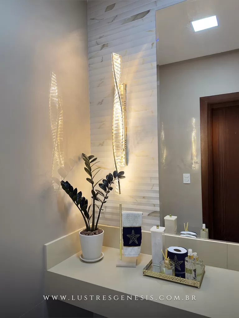 Arandela moderna de cristal com design vertical curvo e iluminação quente, instalada em lavabo elegante com bancada clara, espelho amplo e revestimento em mármore, proporcionando efeito decorativo sofisticado e aconchegante.