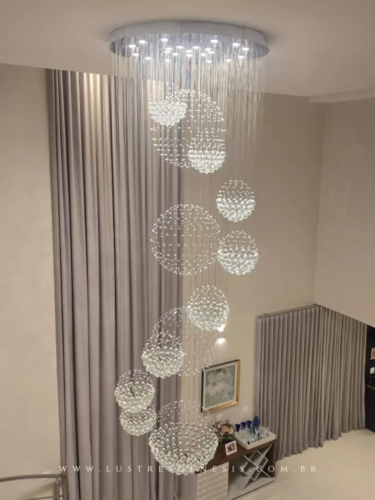 Lustre de cristal para pé-direito duplo com múltiplas esferas suspensas em cascata, iluminando escada em ambiente sofisticado, destacando design moderno e efeito luminoso impactante.