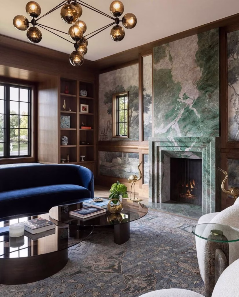 Sala de estar em estilo neo déco com lustre moderno de globos âmbar, paredes revestidas em madeira e painéis artísticos, lareira em mármore verde, sofá curvo em veludo azul e mesa de centro com tampo de vidro.