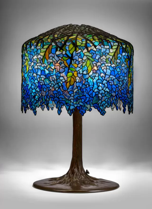 Luminária Tiffany Wisteria com cúpula em vitral azul detalhado com folhas e flores, vidro opalescente em tons de azul e verde, acabamento orgânico com bordas irregulares e base metálica em formato de tronco, ideal para iluminação decorativa clássica e sofisticada.