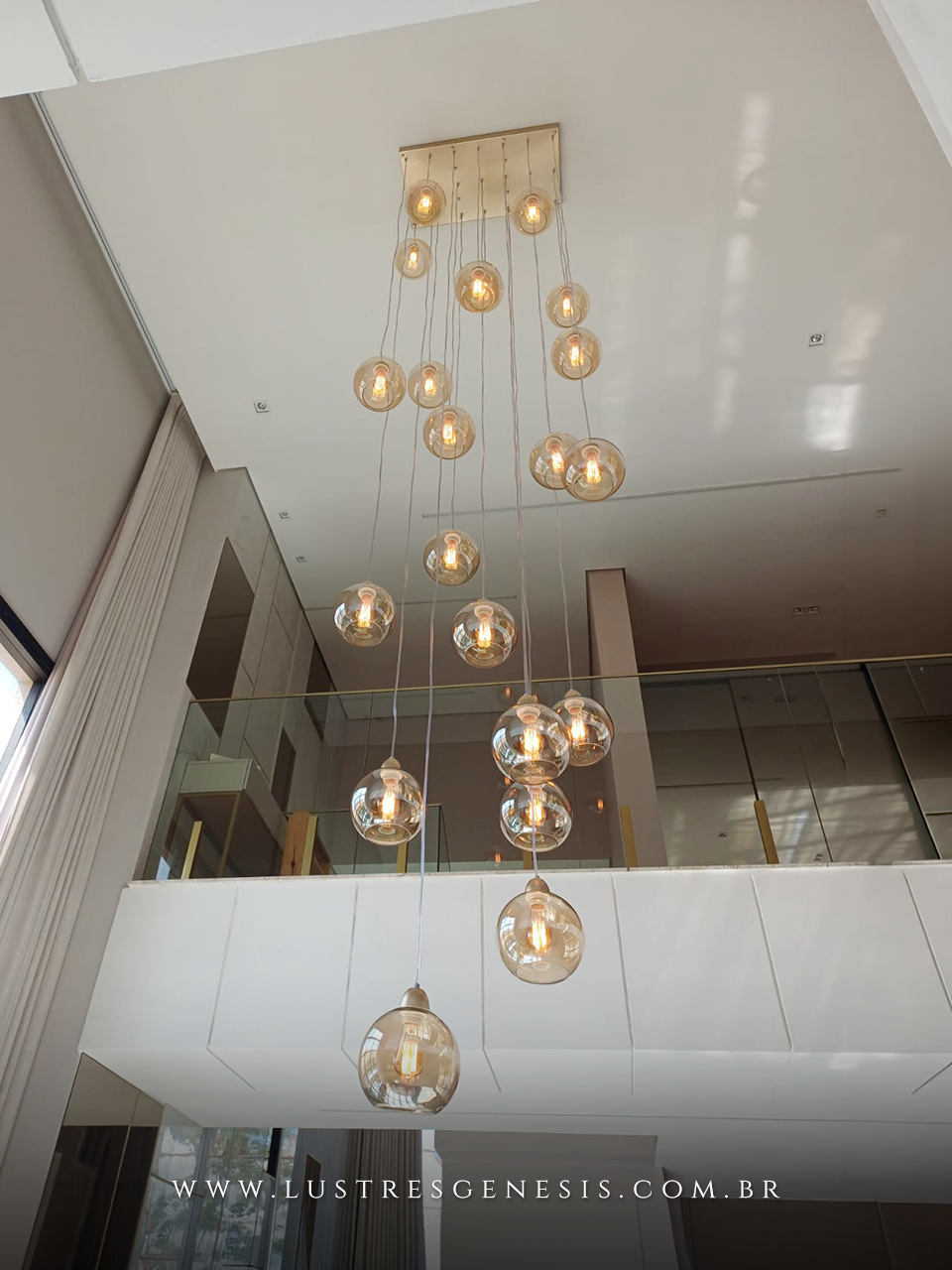 Lustre Sarvah 21 globos da Lustres Gênesis instalado em ambiente com pé direito duplo, com globos de vidro âmbar suspensos em diferentes alturas, criando efeito elegante e sofisticado com iluminação decorativa moderna.