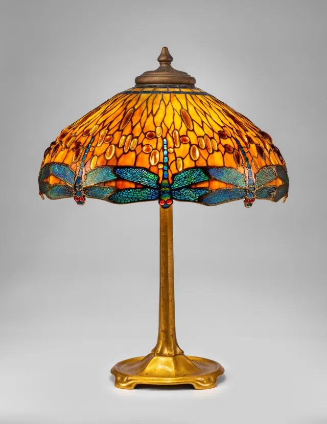 Luminária Tiffany modelo libélula com cúpula em vitral âmbar e detalhes em azul, apresentando insetos estilizados em vidro opalescente, base metálica dourada ornamentada e iluminação decorativa clássica de alto valor artístico.