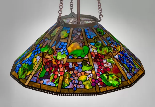 Luminária Tiffany pendente com cúpula em vitral colorido representando cachos de uvas e folhas, vidro opalescente em tons de verde, azul, vermelho e âmbar, estrutura metálica com correntes e iluminação decorativa clássica de alto valor artístico.