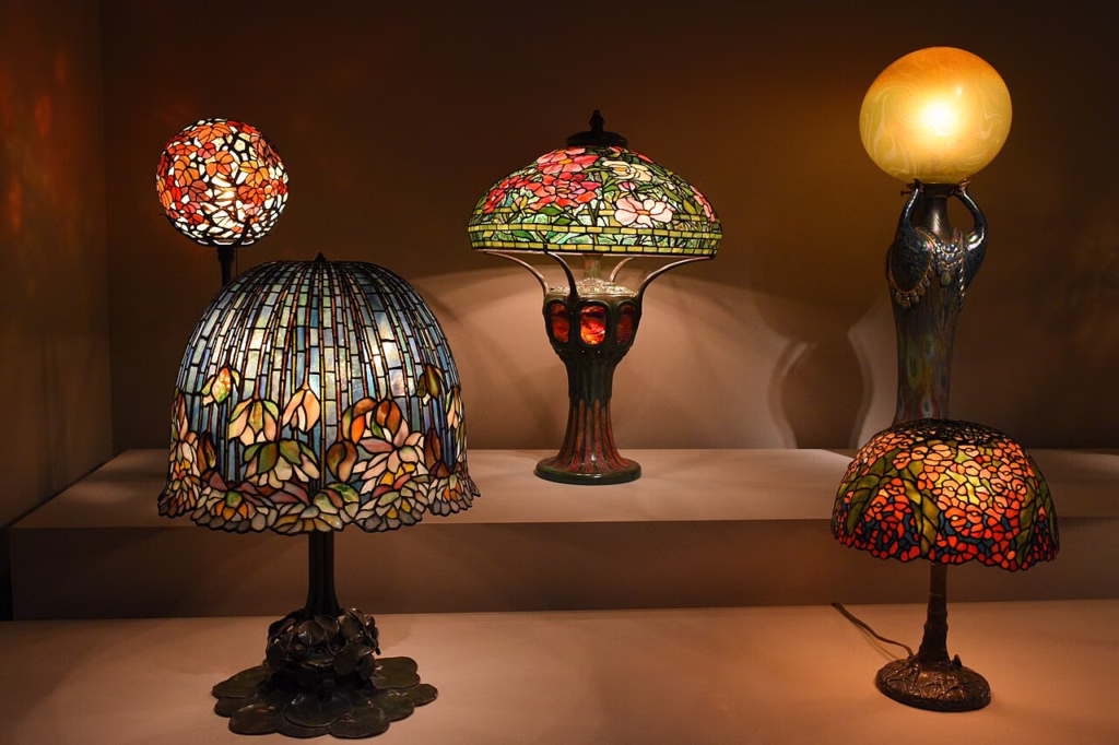 Coleção de luminárias Tiffany expostas no Virginia Museum of Fine Arts, com abajures em vitral colorido e vidro opalescente, apresentando diferentes formatos e padrões florais, bases ornamentadas e iluminação decorativa clássica de alto valor artístico e histórico.