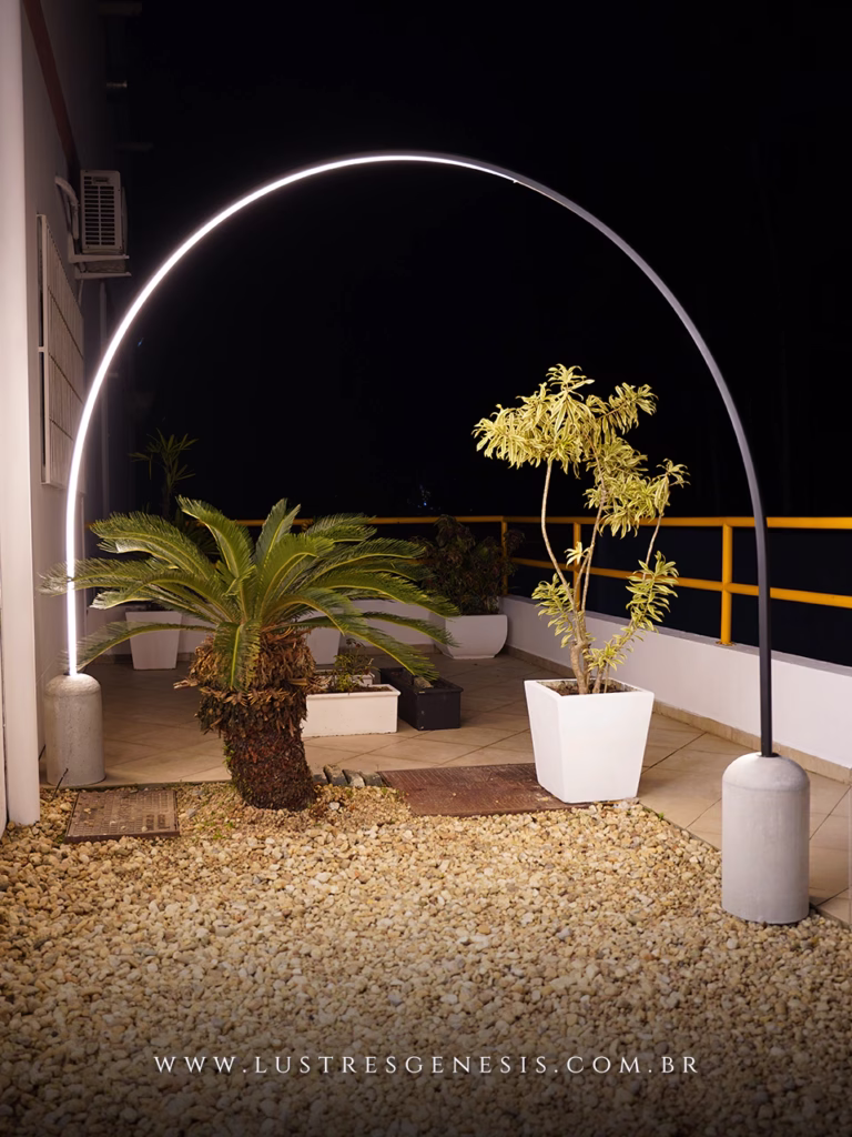 Luminária de piso Arcool com base de concreto em formato de arco iluminado, instalada em área externa decorada com plantas e pedras, destacando design minimalista e sofisticado.