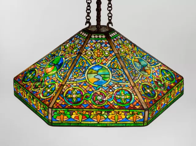Luminária Tiffany pendente com cúpula em vitral geométrico e ornamental, vidro opalescente em cores vibrantes como azul, verde, vermelho e âmbar, com padrões circulares e detalhes simétricos, estrutura metálica com correntes e iluminação decorativa clássica.