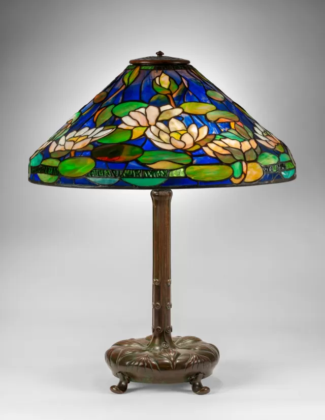 Luminária Tiffany modelo lírio-d’água com cúpula em vitral colorido em tons de azul, verde e branco, apresentando flores e folhas sobre a água, vidro opalescente detalhado e base metálica ornamentada, ideal para iluminação decorativa clássica e sofisticada.