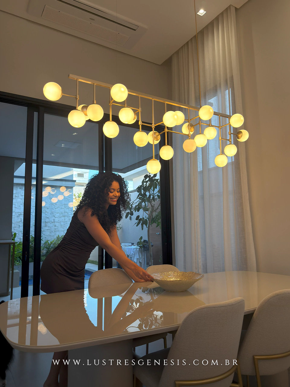 Luminária de teto pendente Cacho Jabuticaba com 23 globos leitosos instalada sobre mesa de jantar moderna, com a Stefanny Borges arrumando a mesa em ambiente sofisticado.