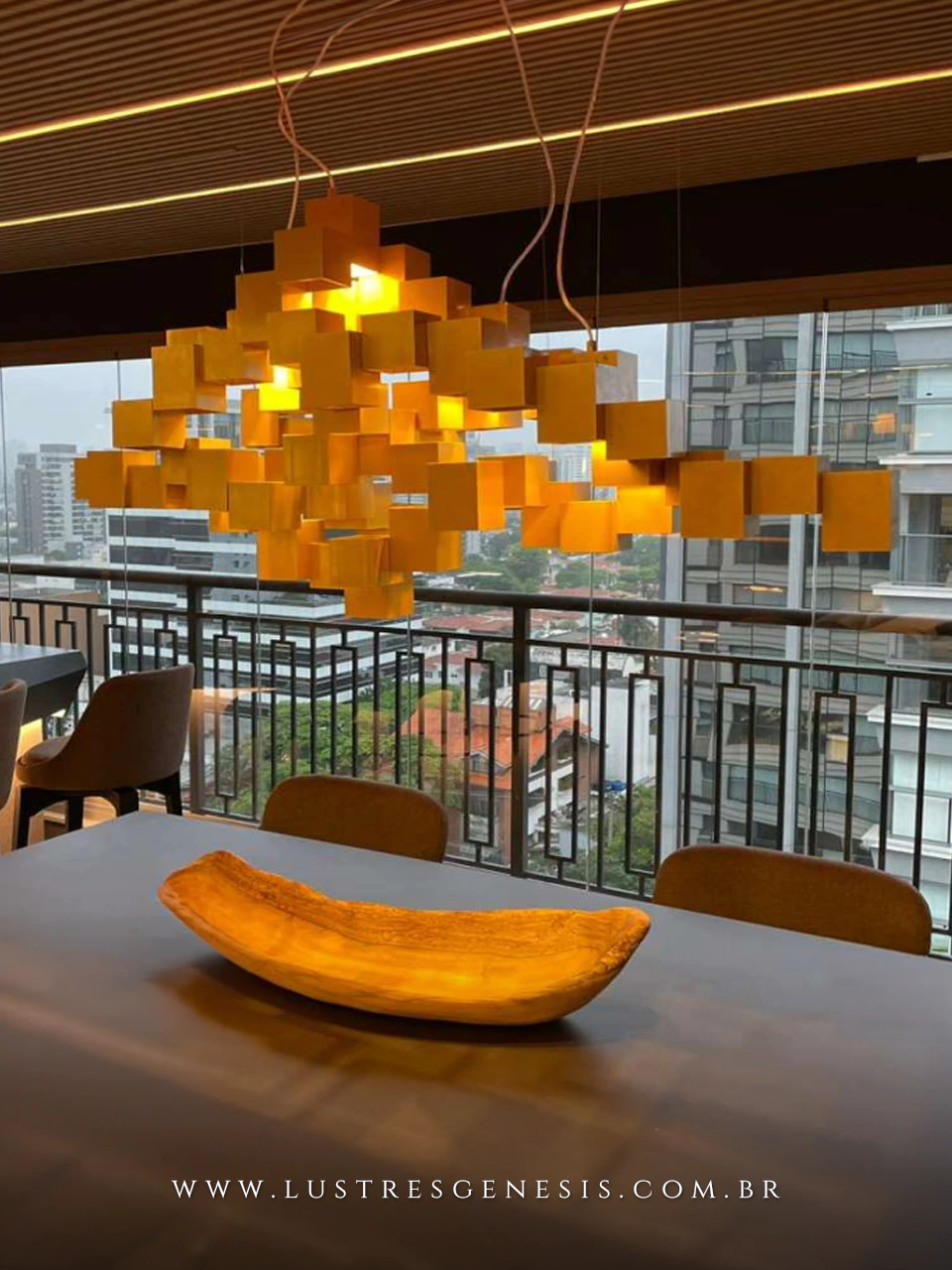 Lustre pendente cubos suspensos arte dourado LED iluminando sala de jantar moderna com mesa e cadeiras, vista para varanda e cidade ao fundo.