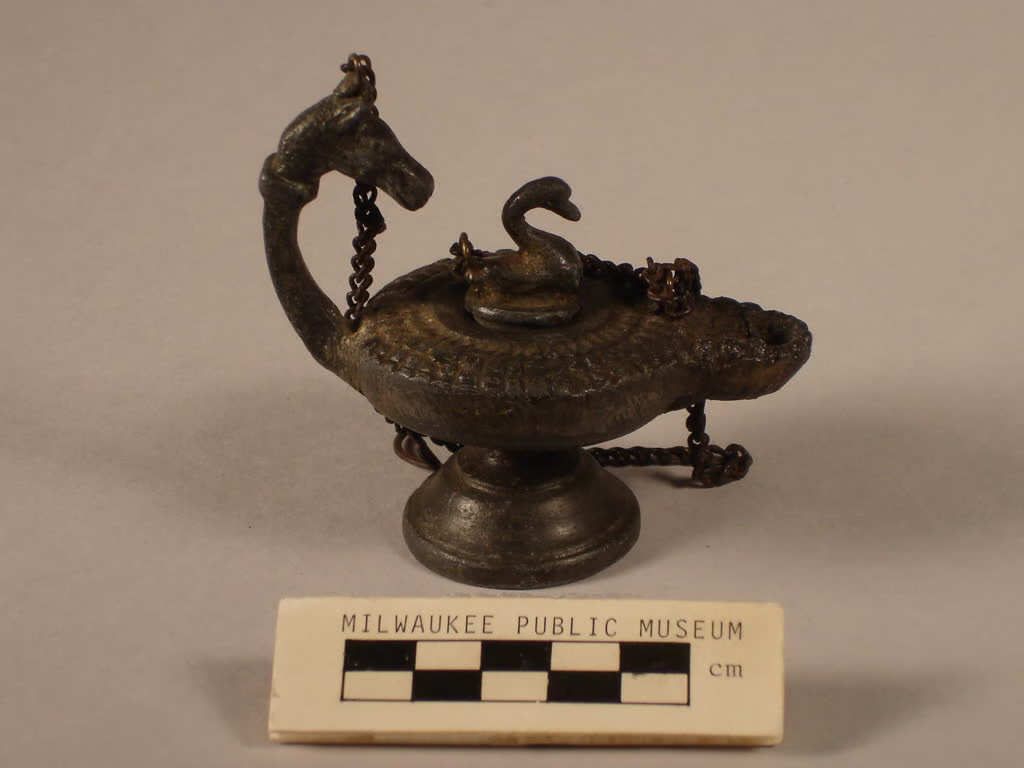 Lamparina antiga de metal com formato ornamental inspirado em um cisne, apoiada sobre base elevada. A peça possui reservatório para óleo, tampa decorativa, bico para pavio e alça arqueada em formato de pescoço de ave, representando a evolução das luminárias utilitárias para modelos com valor artístico e decorativo.