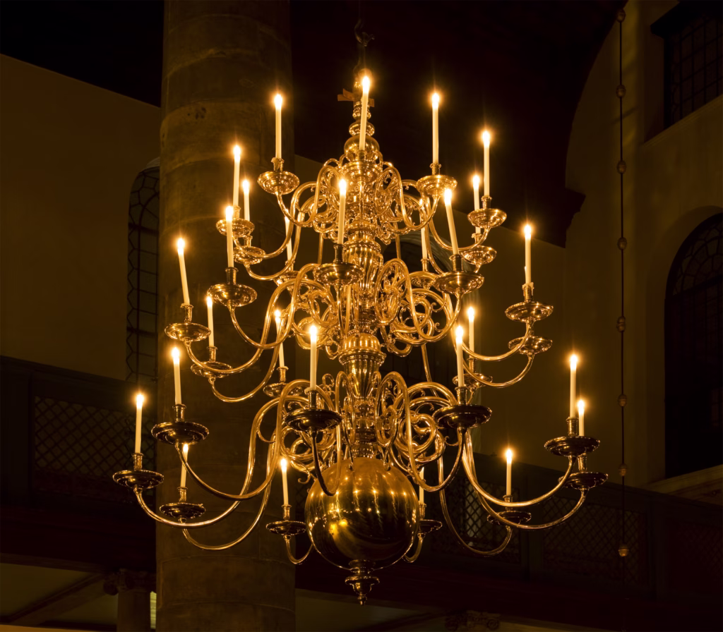 Lustre de velas suspenso em ambiente histórico, com múltiplos braços metálicos ornamentados sustentando dezenas de velas acesas. A peça produz iluminação quente e acolhedora, representando os primeiros lustres utilizados em igrejas, castelos e grandes salões antes da eletrificação.