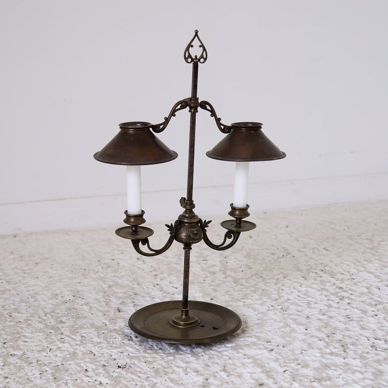 Luminária de mesa antiga em bronze com duas velas protegidas por pequenas cúpulas metálicas. A peça possui braços ornamentados, base circular e detalhes decorativos clássicos, representando a transição entre os candelabros tradicionais e as primeiras luminárias de mesa mais sofisticadas.