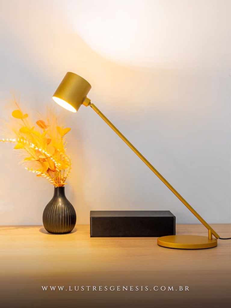 Luminária de mesa abajur Joy dourada acesa sobre escrivaninha, ao lado de vaso preto decorativo e caixa preta, ideal para leitura em quartos, escritórios e salas.