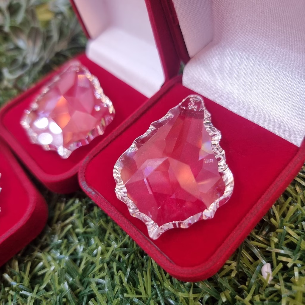 Pingente de cristal Asfour lapidado em formato de folha, com facetas que refletem a luz, exibido em um estojo de veludo vermelho. A peça é utilizada como cristal decorativo em lustres clássicos e contemporâneos, proporcionando brilho e sofisticação à iluminação.