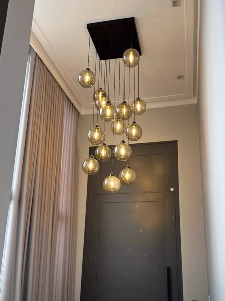 Lustre pendente Sarvah com 15 globos de vidro e base quadrada instalado em hall de entrada com pé direito duplo, iluminação moderna e elegante para ambientes altos.